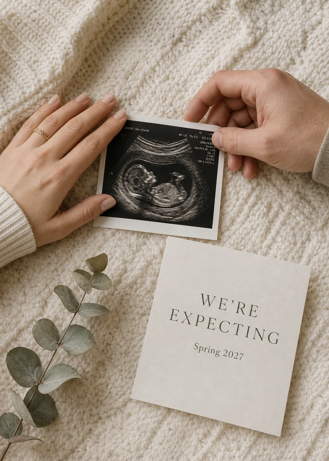 Overhead flat-lay pregnancy announcement: a sonogram framed by two hands beside a We're Expecting due-date card on a cream knit blanket.