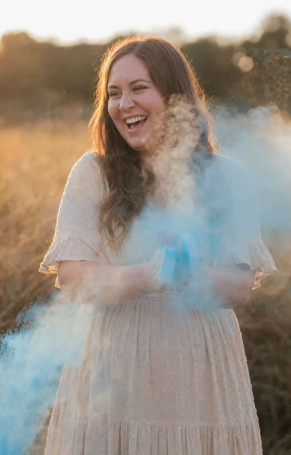 A laughing parent-to-be in a golden-hour field as a soft cloud of pale blue powder bursts out in front of them.