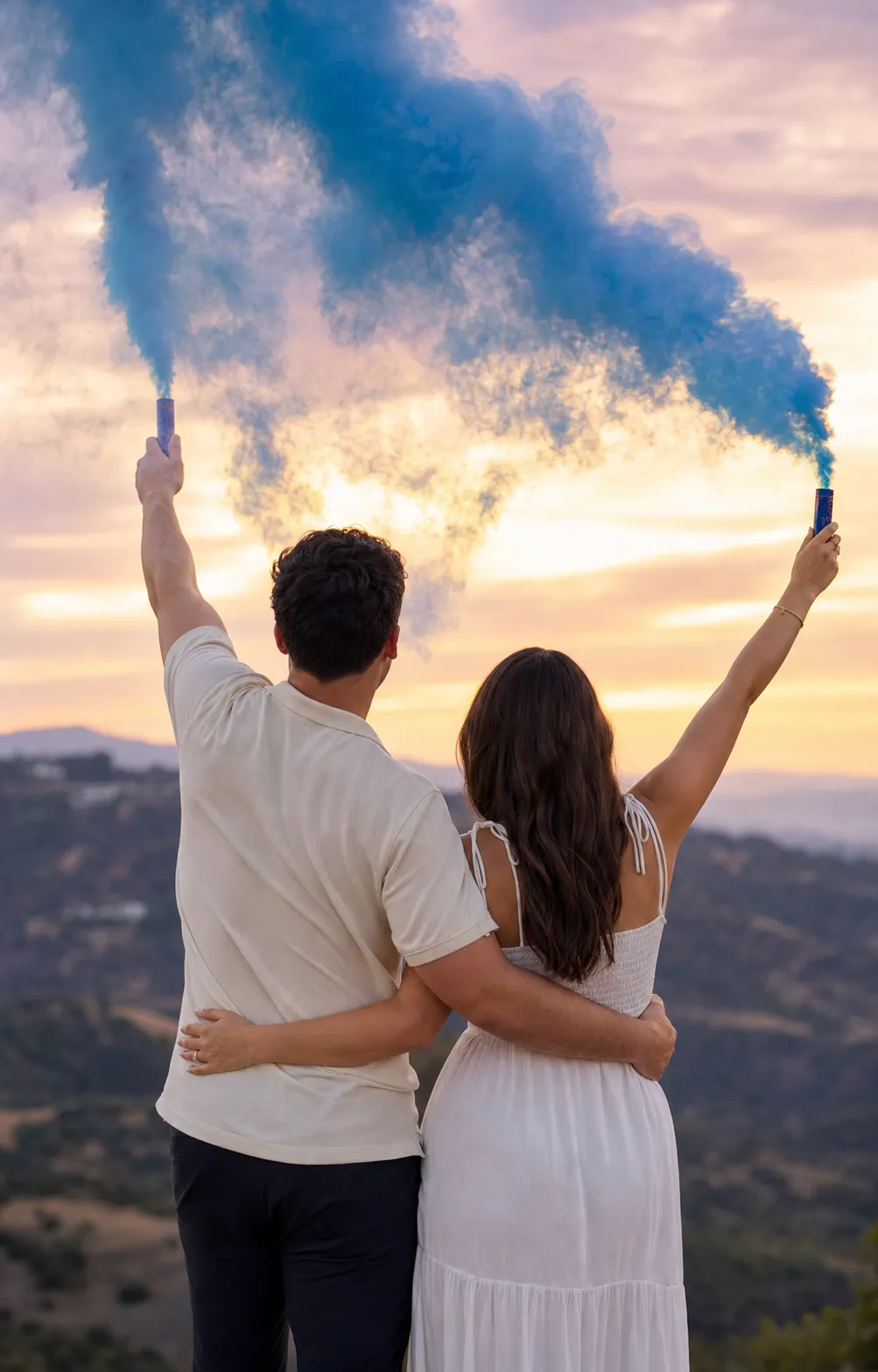 A couple seen from behind on a dusk hillside raising smoke sticks as a cloud of soft blue colored smoke pours up against the sky.