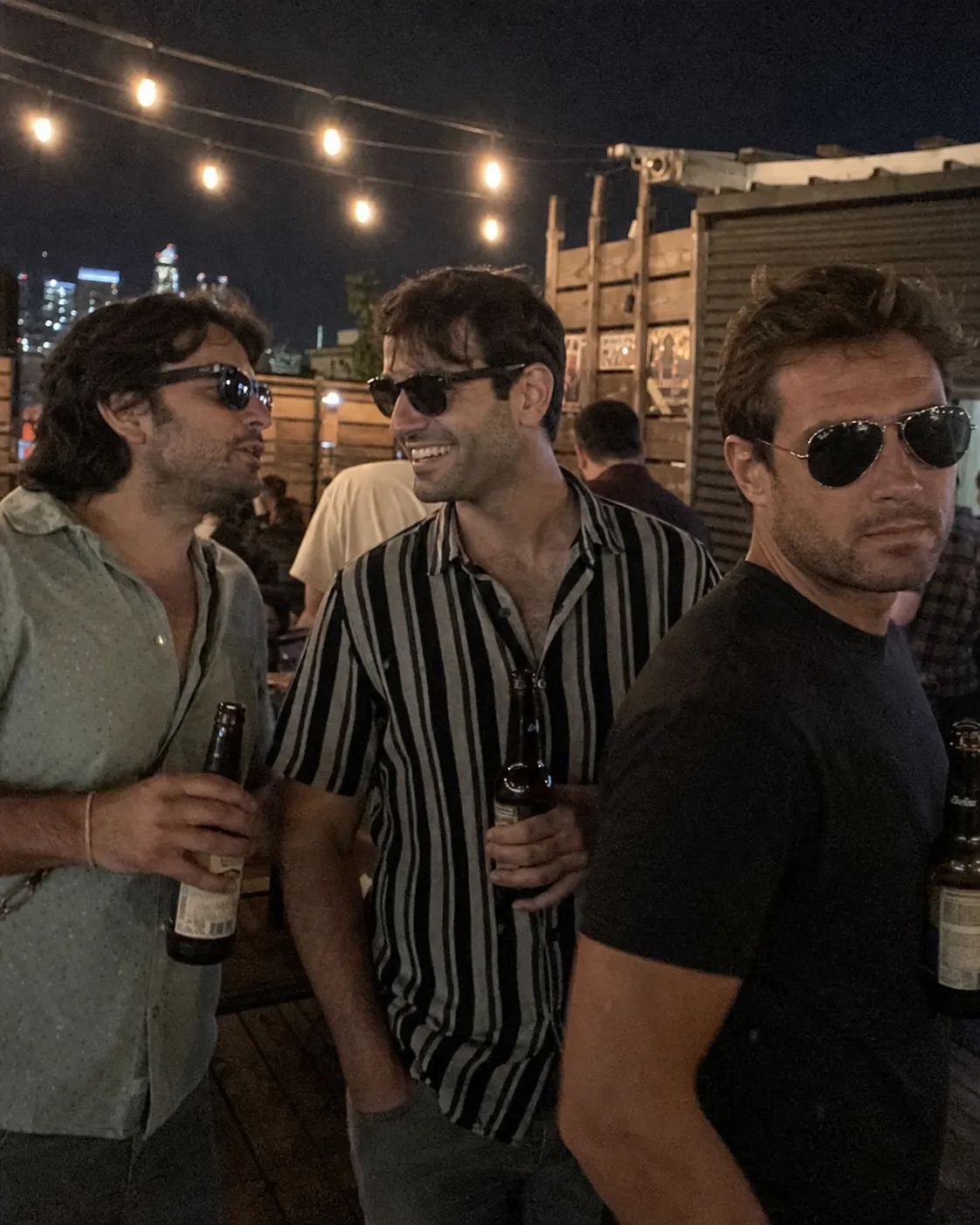 A photoreal 4:5 vertical dating-app lead photo of the same 32-year-old man standing in a group of three friends at a dim rooftop bar at night, all wearing dark sunglasses and holding beers, his face occupying about 25% of the vertical frame partially turned from the camera, the compound failure case.