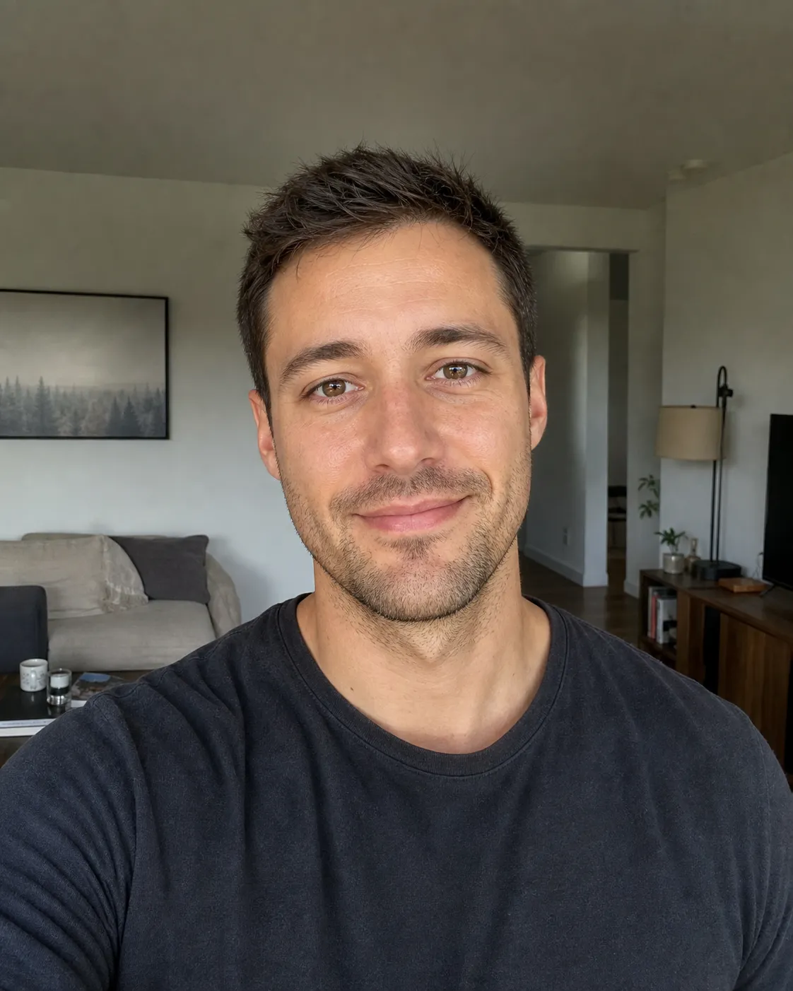 A photoreal vertical dating-profile photo of a 31-year-old man with short dark-brown hair and light stubble, his face centered and occupying about 65 percent of the frame with clear headroom above his head and his shoulders visible, a warm half-smile and direct eye contact, soft natural window light from the front-left, a plain charcoal tee, a softly blurred home interior behind him, the zoom-safe lead that survives Tinder's deck crop.