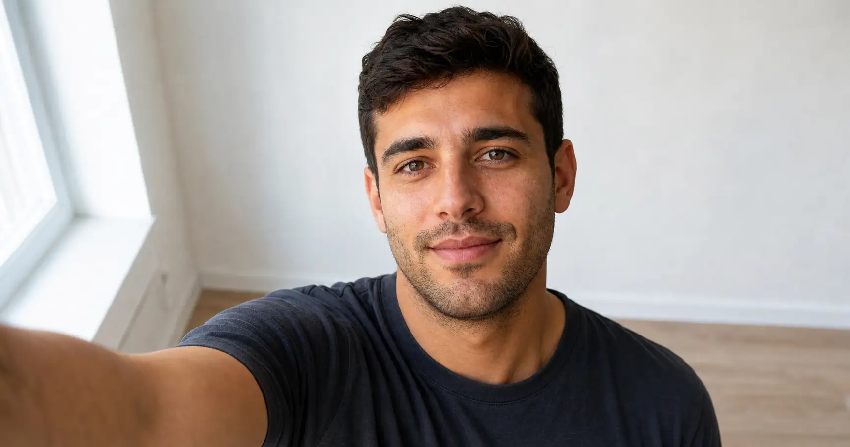 A man taking a flattering selfie with the phone held just above eye level and soft window light on his face.