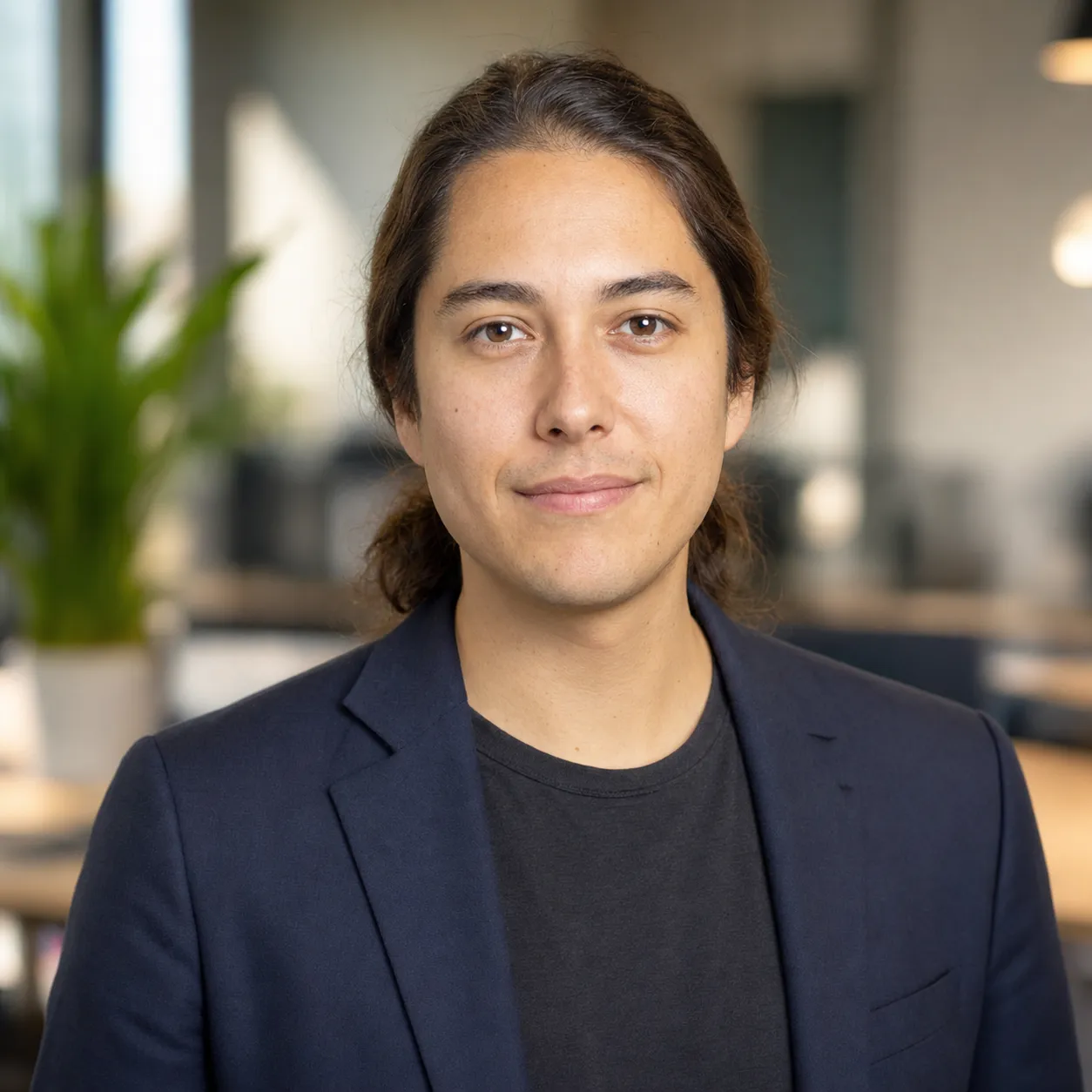 LinkedIn headshot example for a tech individual contributor, soft-shoulder navy blazer relaxed over a charcoal cotton tee against a warm soft-bokeh office backdrop, soft balanced front-light, identity-locked 1:1 square recruiter-screen format.
