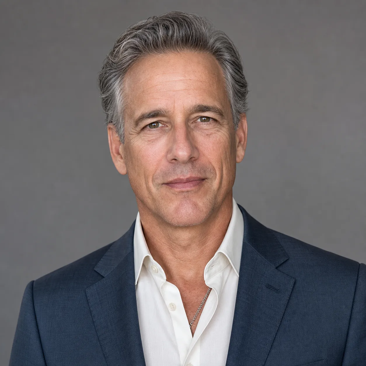 LinkedIn headshot example for a 50+ executive, refined slate-blue tailored blazer over a cream silk shirt against a soft mid-gray studio backdrop, soft balanced front-light, age-appropriate texture preserved, identity-locked 1:1 square recruiter-screen format.