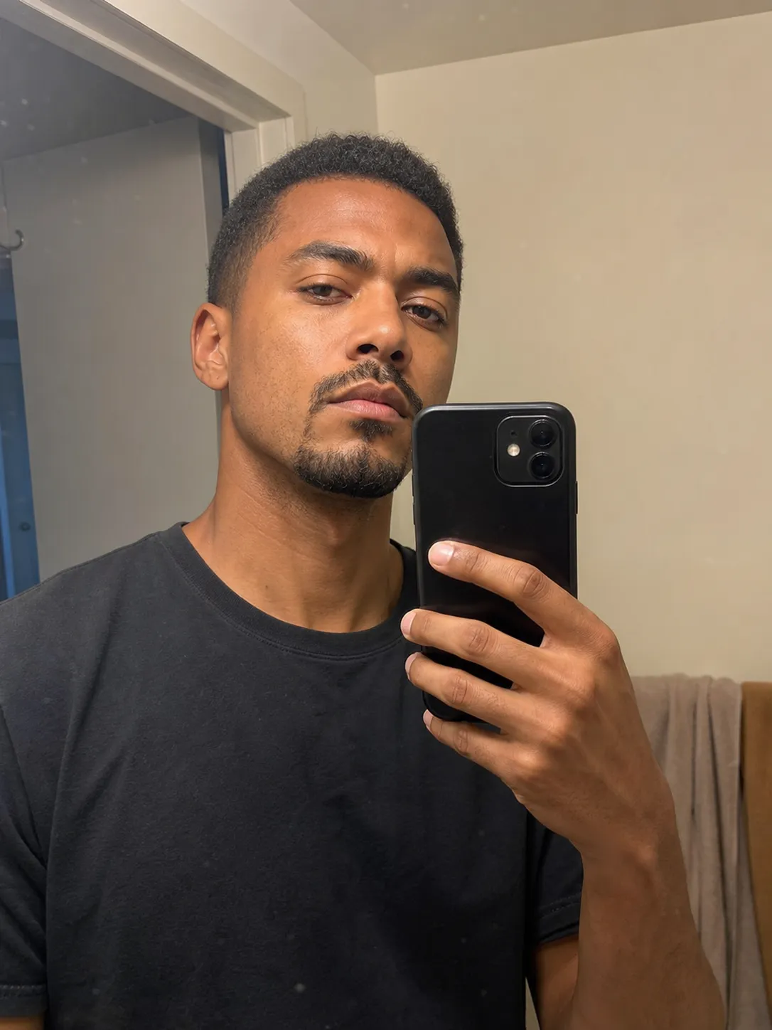 A plain indoor mirror selfie of a man in a dark t-shirt, before the AI photoshoot.