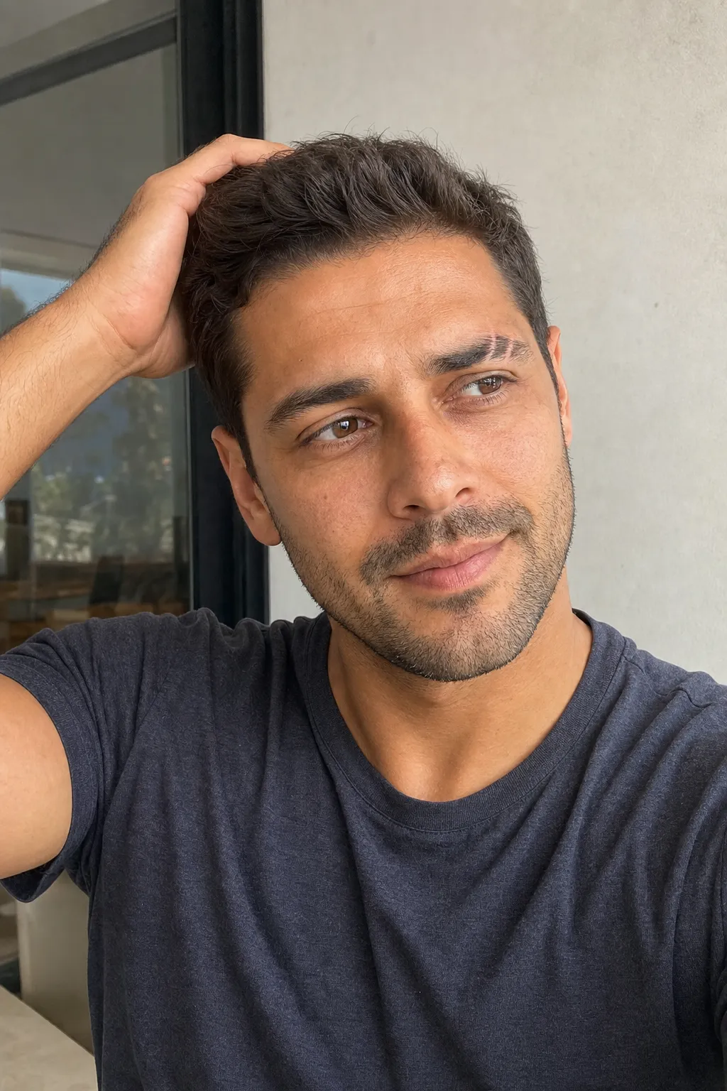 A man taking a candid-feeling selfie looking off to the side with one hand running back through his hair.