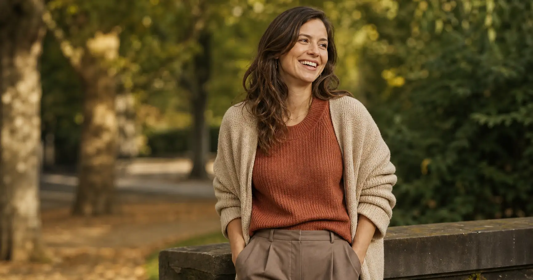 A relaxed everyday woman in a coordinated warm-neutral outfit, a rust knit top with an open cream cardigan and taupe trousers in one palette, half-laughing in soft natural light against a softly blurred park background.