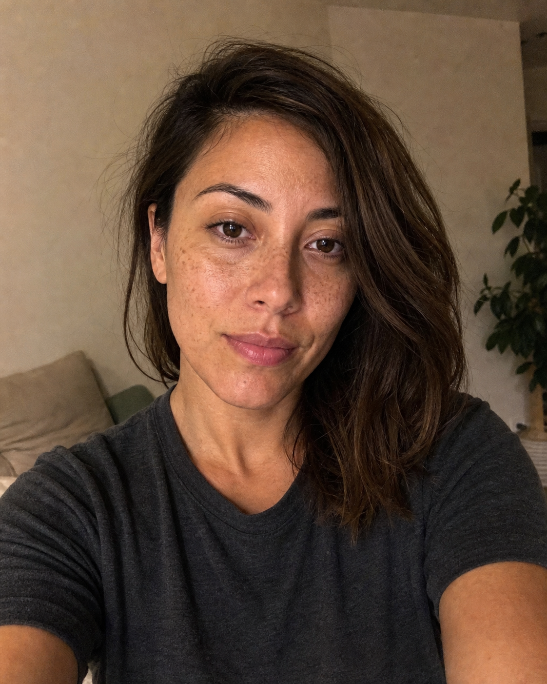 The kind of input the 1-selfie identity-anchor workflow runs against: a casual at-home iPhone selfie of a 30-year-old woman with shoulder-length brown hair, slight freckles across the cheekbones and nose bridge, a plain dark grey crew-neck t-shirt, a faint closed-mouth smile, and mediocre overhead warm-bulb indoor lighting, deliberately the kind of single decent selfie a reader would find in their own camera roll rather than a polished dating-app photo.