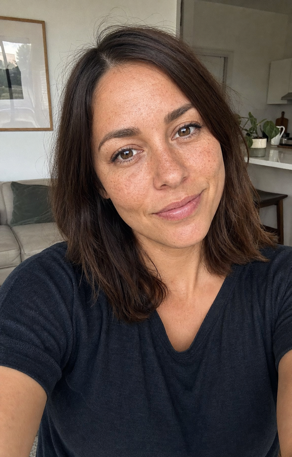 The AFTER side of the FaceTune-to-real downgrade comparison: the same 30-year-old woman from the BEFORE image but rendered with the downgrade rule applied, slight freckles on cheekbones visible and preserved, no cheek or chin contour smoothing, natural facial micro-asymmetry preserved, soft natural window light replacing the ring-light flatness, phone-held casual selfie in a real lived-in living-room corner rather than a bathroom mirror, identity-locked 4:5 vertical dating-app lead-photo format demonstrating what an AI render looks like when the FaceTune-to-real paste-line is in the rules block.