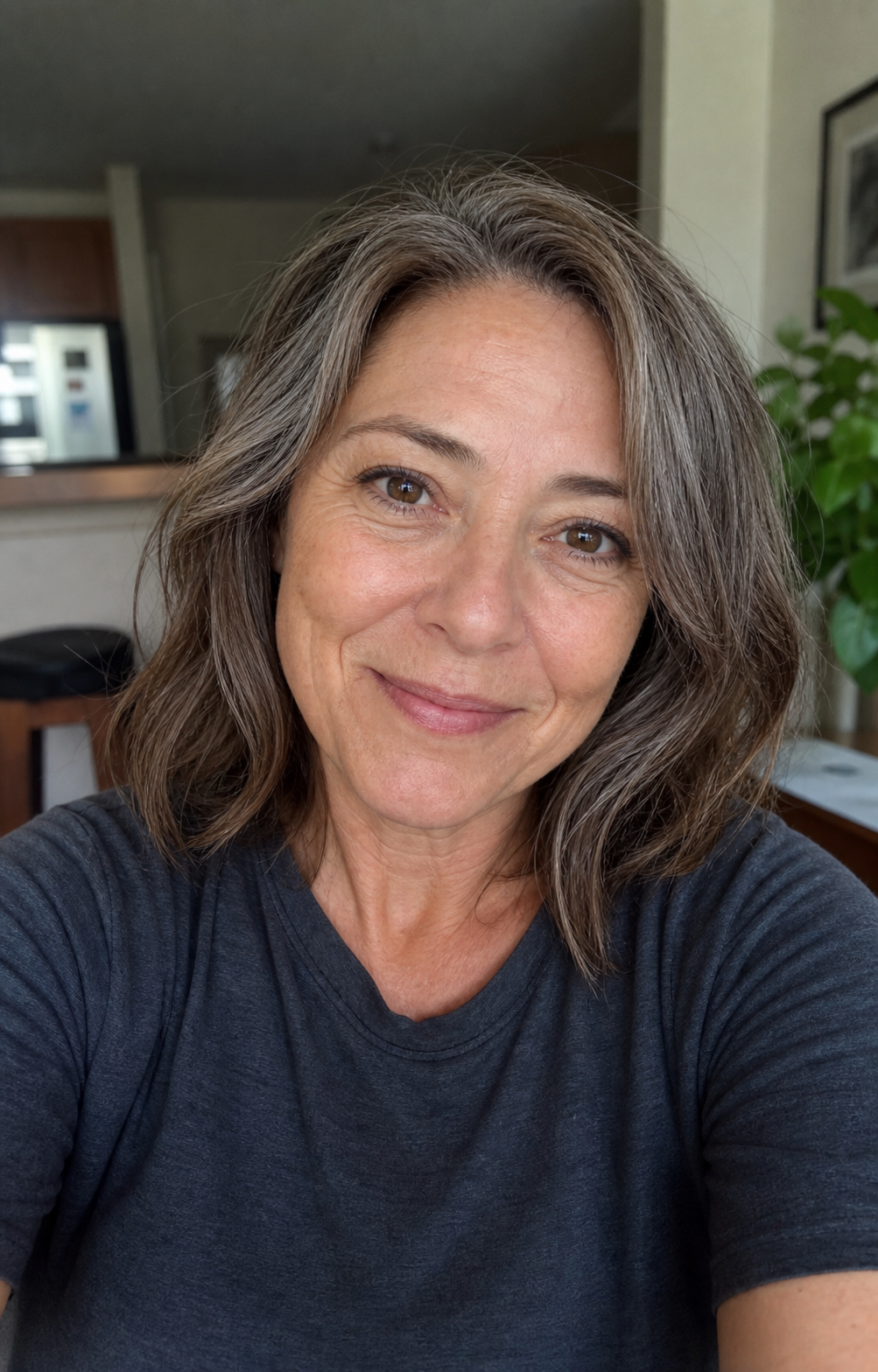 Close-up candid selfie of the same 50-year-old woman from the hero pack, showing soft natural window light, a warm half-smile that crinkles the eyes, grey-streaked shoulder-length hair, visible nasolabial lines, and mid-50s skin texture — the lead-photo slot of an over-40 dating profile rendered with the aging-preservation rule held.