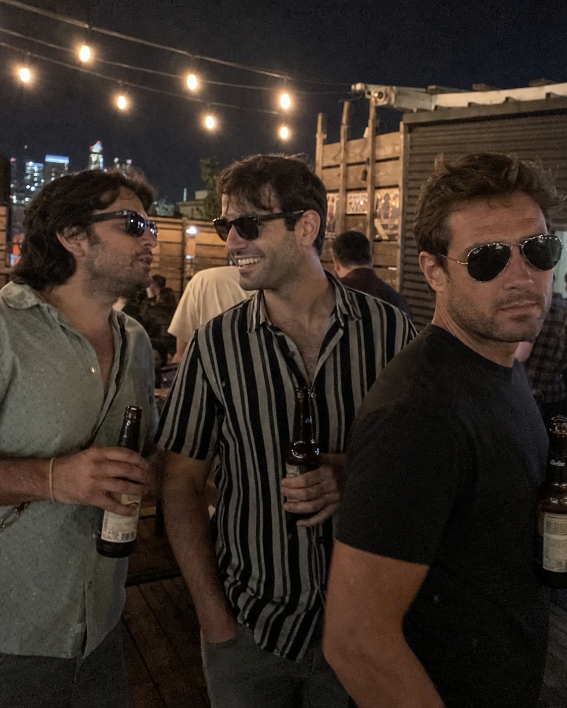 A photoreal 4:5 vertical dating-app lead photo of the same 32-year-old man standing in a group of three friends at a dim rooftop bar at night, all wearing dark sunglasses and holding beers, his face occupying about 25% of the vertical frame partially turned from the camera, the compound failure case.