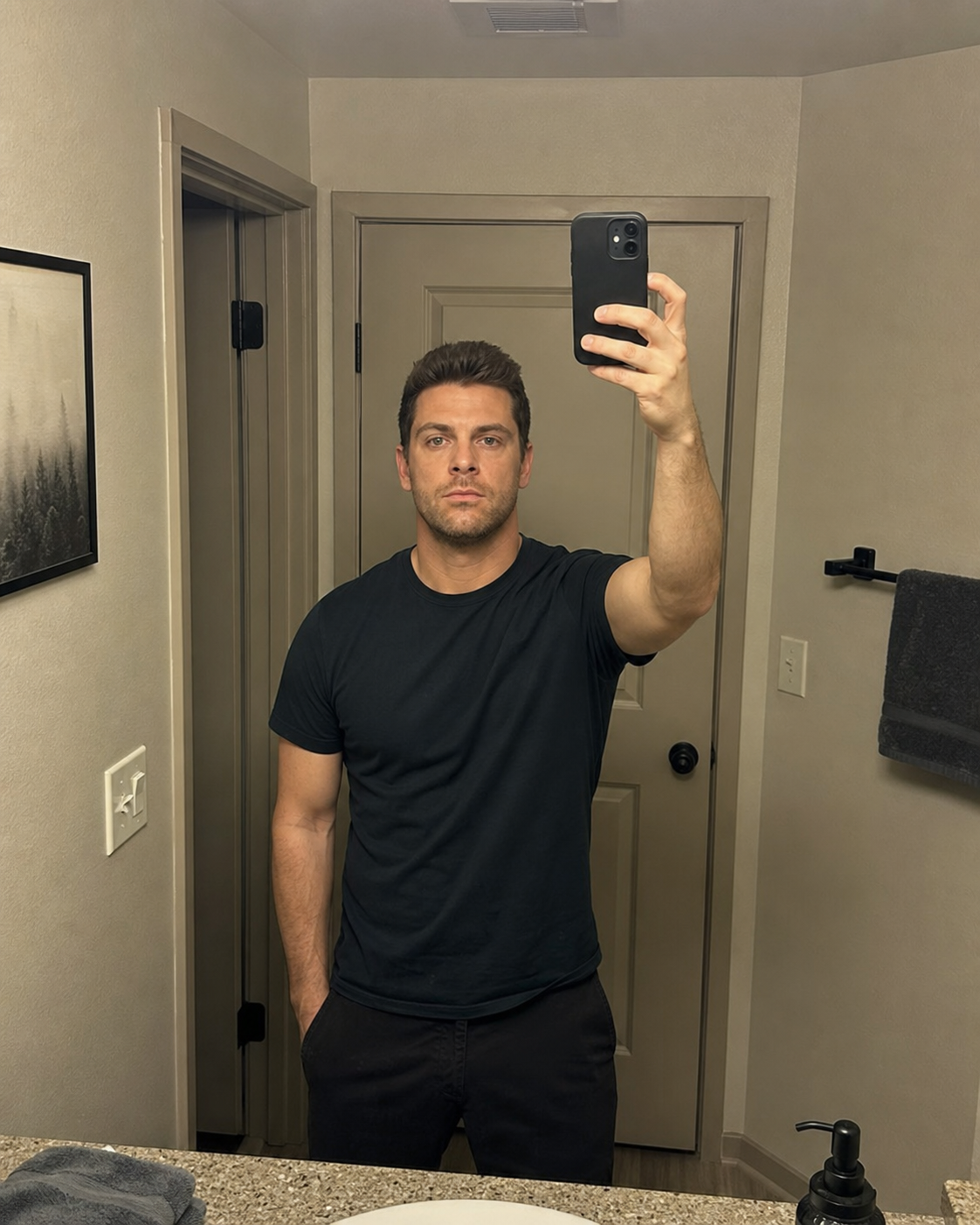 A photoreal 4:5 vertical dating-app lead photo of the same 32-year-old man taken in a bathroom mirror at arm's length under harsh fluorescent overhead light, his face occupying only about 40% of the vertical frame and his eyes landing at the dead-center rather than the upper-third grid line, the compound rule-break common to the bathroom-mirror selfie canon.