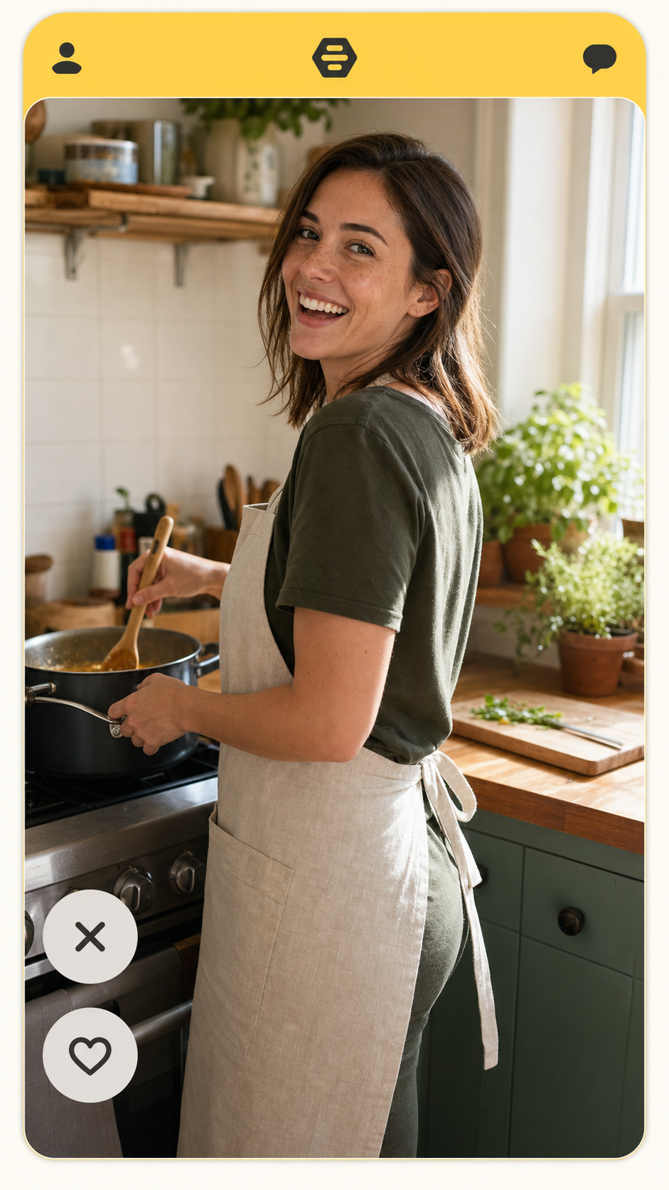 Bumble lead-photo example: a 9:16 full-body shot of a woman around thirty mid-laugh while mid-stir at a sunlit kitchen counter, identity-locked, cream linen apron over a dark olive tee, freckles on the cheekbones visible, displayed inside a Bumble profile card with a soft yellow accent bar, demonstrating the Bumble conversation-opener lead-photo move in 2026.