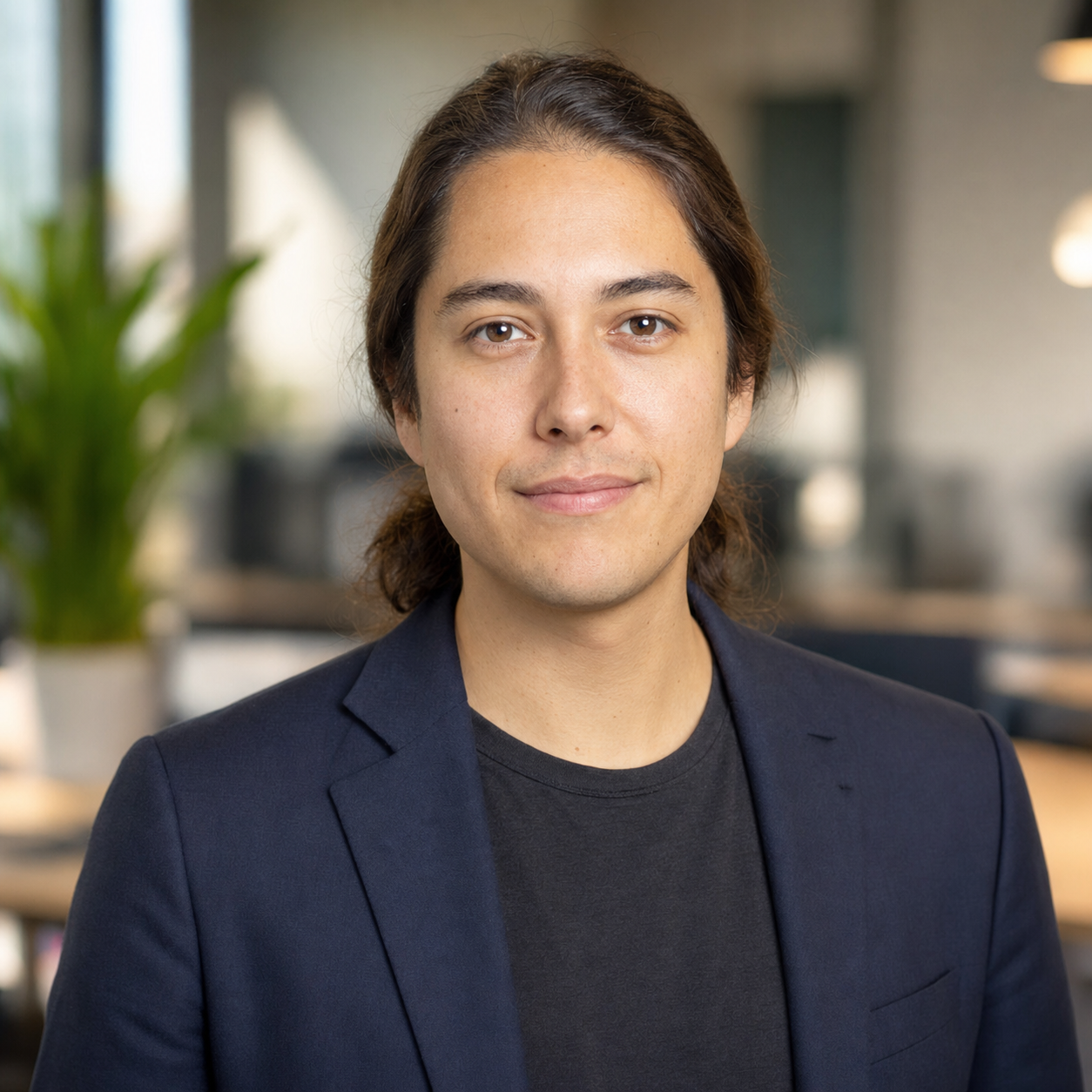 LinkedIn headshot example for a tech individual contributor, soft-shoulder navy blazer relaxed over a charcoal cotton tee against a warm soft-bokeh office backdrop, soft balanced front-light, identity-locked 1:1 square recruiter-screen format.