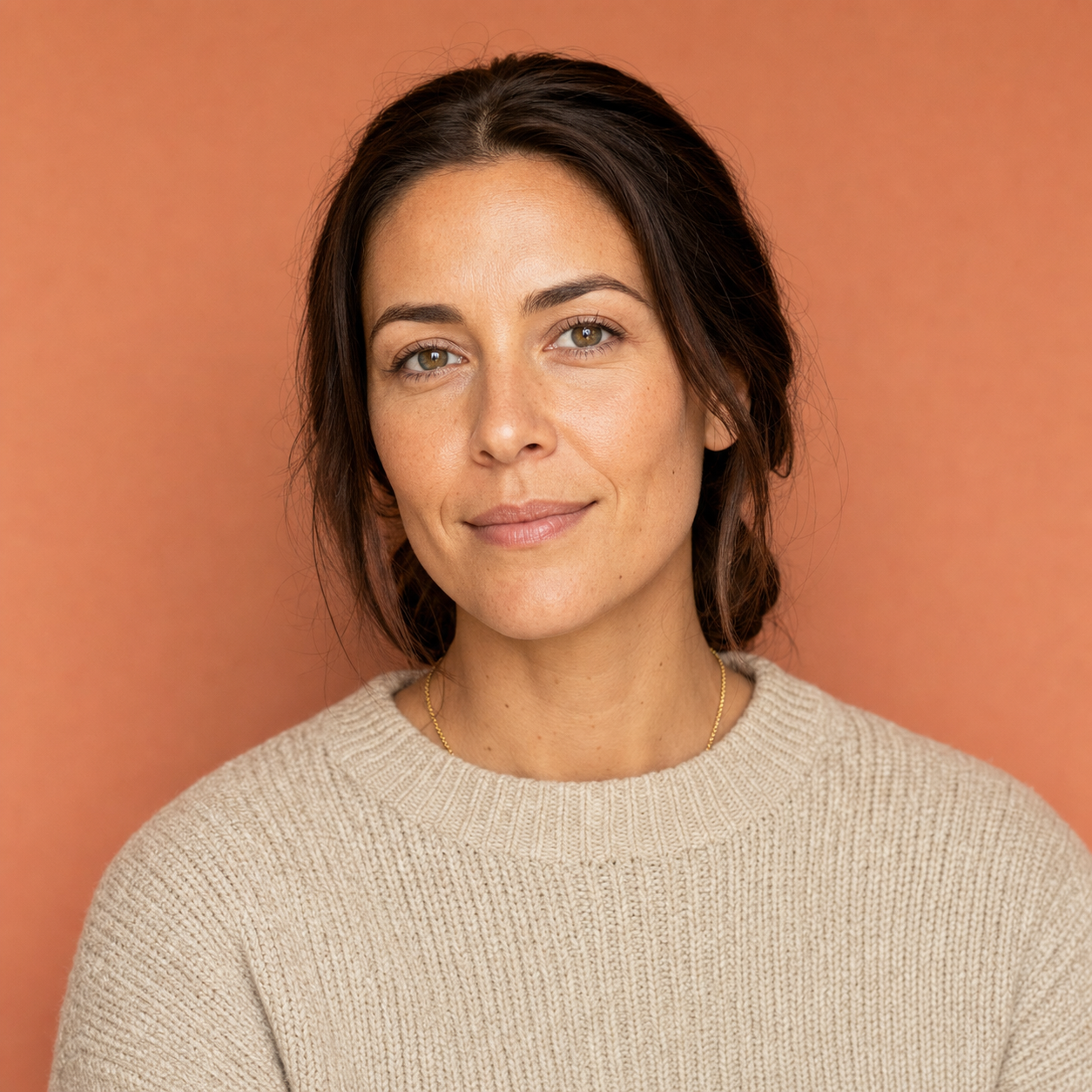 LinkedIn headshot example for a creative-industry professional, oatmeal heavyweight knit sweater with a thin gold chain against a soft warm-terracotta painted wall, soft balanced front-light, identity-locked 1:1 square recruiter-screen format.