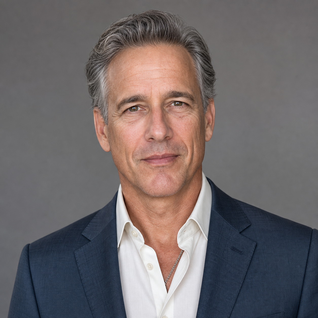 LinkedIn headshot example for a 50+ executive, refined slate-blue tailored blazer over a cream silk shirt against a soft mid-gray studio backdrop, soft balanced front-light, age-appropriate texture preserved, identity-locked 1:1 square recruiter-screen format.